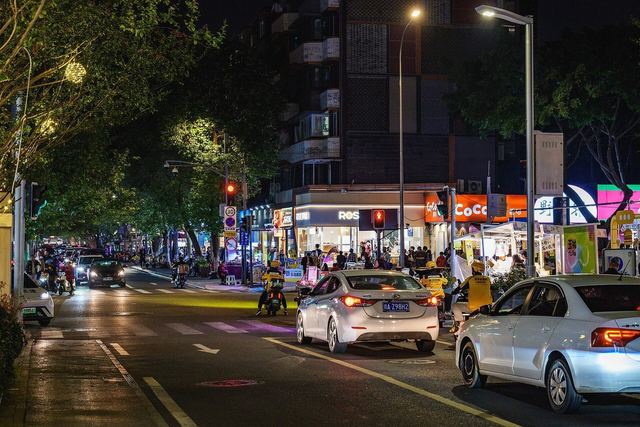 Cars and scooters waiting at a lit crossroads in Chengdu's Yulin at night, shopfronts glowing